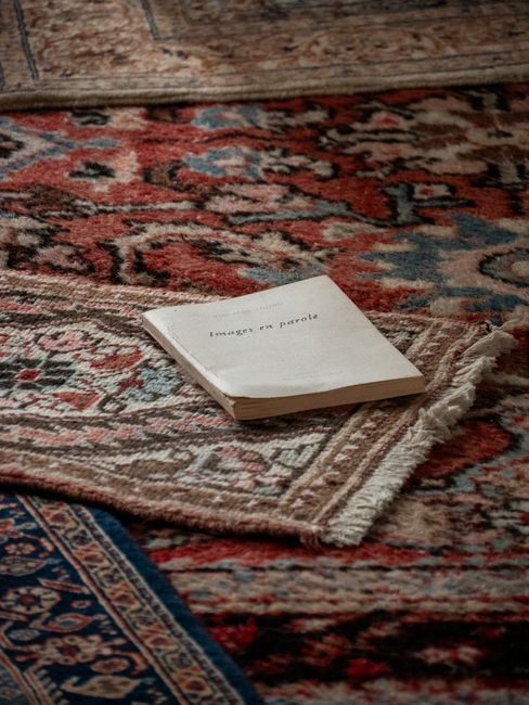 A professional cleaner dressed in white protective clothing, including a face mask, gloves, and shoe covers, is using a vacuum or steam cleaning tool on a beige, textured carpet in a well-lit living r