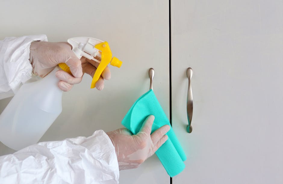 A person wearing white cleaning gloves and a protective gown is holding a white spray bottle with a yellow nozzle, applying a cleaning solution to a turquoise cloth. The cloth is used to wipe the surf