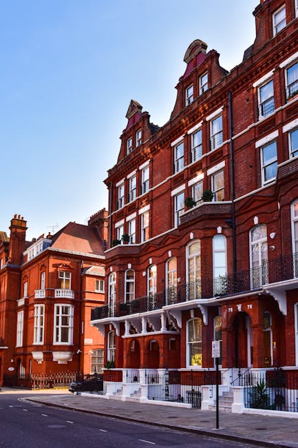A row of Victorian-style terraced houses with brick facades and white window frames, situated behind a large green park with well-maintained grassy lawns and a few scattered trees. The park appears cl