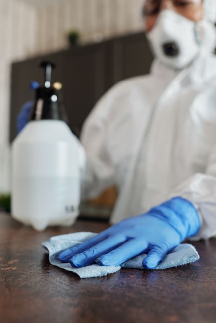 A professional cleaner from Hammersmith Cleaners is engaged in surface cleaning within a domestic or commercial setting, dressed in full protective gear including a white coverall, face mask, goggles,